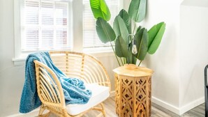 A wicker chair with a blue blanket, a wooden side table with a plant, and a window with blinds. - OKC 9 · Charming Carriage Cottage from 1920`s! (Oklahoma City)