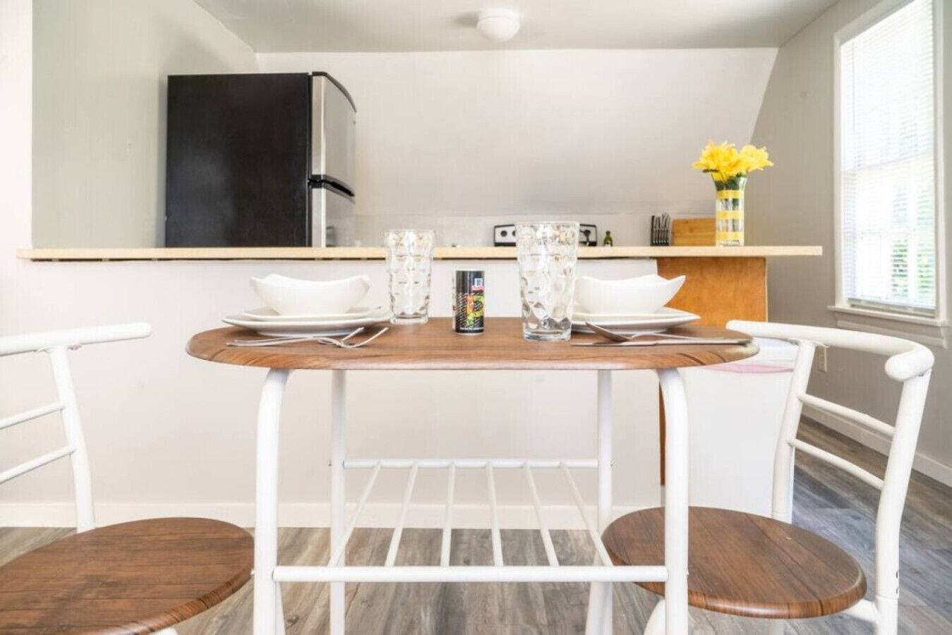 Une petite cuisine équipée d’un réfrigérateur, d’une table en bois dressée pour deux personnes et d’une fenêtre donnant sur l’extérieur.