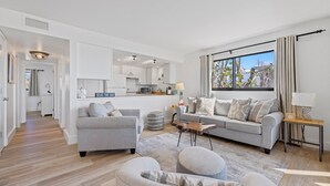 Moderni olohuone, jossa on harmaa sohva, rahi ja sohvapöytä. Taustalla näkyy keittiö, jossa on valkoiset kaapit ja mikroaaltouuni.