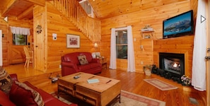 A cozy log cabin interior with a fireplace, wooden walls, a red sofa, a wooden coffee table, and a television mounted above the fireplace.