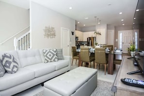 Un salon moderne comprenant un canapé, un pouf, une table à manger, des chaises, un coin cuisine et un escalier.