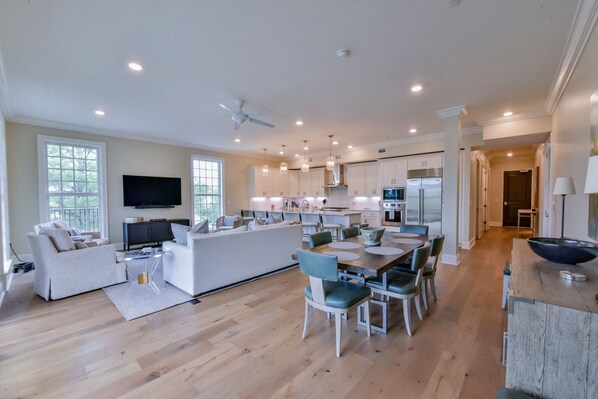 A spacious living area with a sofa, coffee table, and TV. There is a dining area with a table and chairs. The kitchen has white cabinets and a large island.