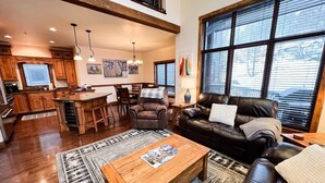 A spacious living area with a leather sofa, wooden coffee table, and a large window with blinds. - Luxury Home | Hot Tub | Ski In / Ski Out | Warmers (Whitefish)
