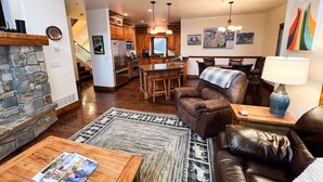 A living room with a fireplace, a stone wall, a wooden coffee table, a leather sofa, and a dining area with a table and chairs. - Luxury Home | Hot Tub | Ski In / Ski Out | Warmers (Whitefish)