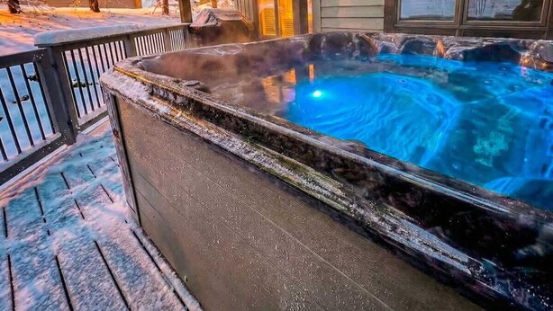 A hot tub on a wooden deck with a view of snow-covered trees and a building in the background. - Luxury Home | Hot Tub | Ski In / Ski Out | Warmers (Whitefish)