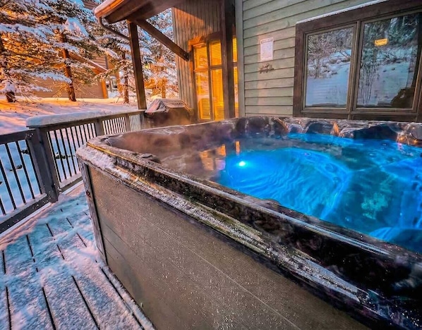 A hot tub on a wooden deck with a view of snow-covered trees and a building in the background.