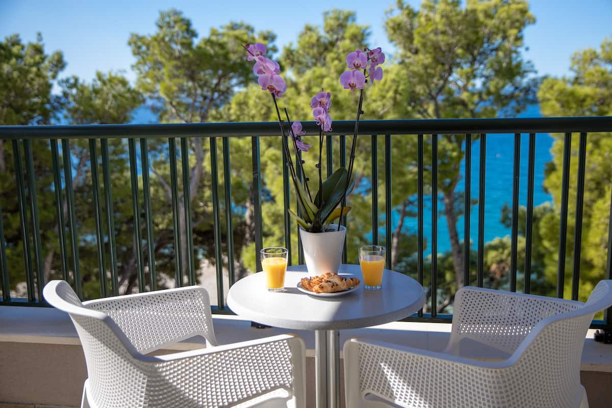 A balcony with white wicker chairs, a small round table with a potted plant, two glasses of orange juice, and a plate of pastries.