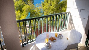 Un balcón con una mesa dispuesta para el desayuno, que incluye cruasanes, café y zumo de naranja, con vistas panorámicas al mar y a los árboles.