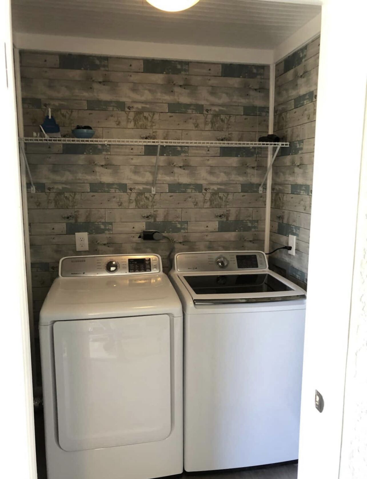 A laundry room with two white front-loading washers and dryers, a metal shelf above, and a wooden wall with a distressed finish.