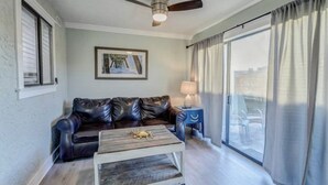 Una sala de estar con un sofá de cuero, una mesa de centro de madera, un ventilador de techo, un cuadro enmarcado en la pared, una lámpara y un amplio ventanal con cortinas.