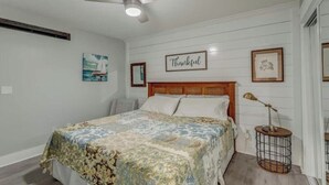 Un dormitorio con cama, ventilador de techo, letrero en marco que dice 'Thankful' y una pequeña mesa redonda con lámpara.