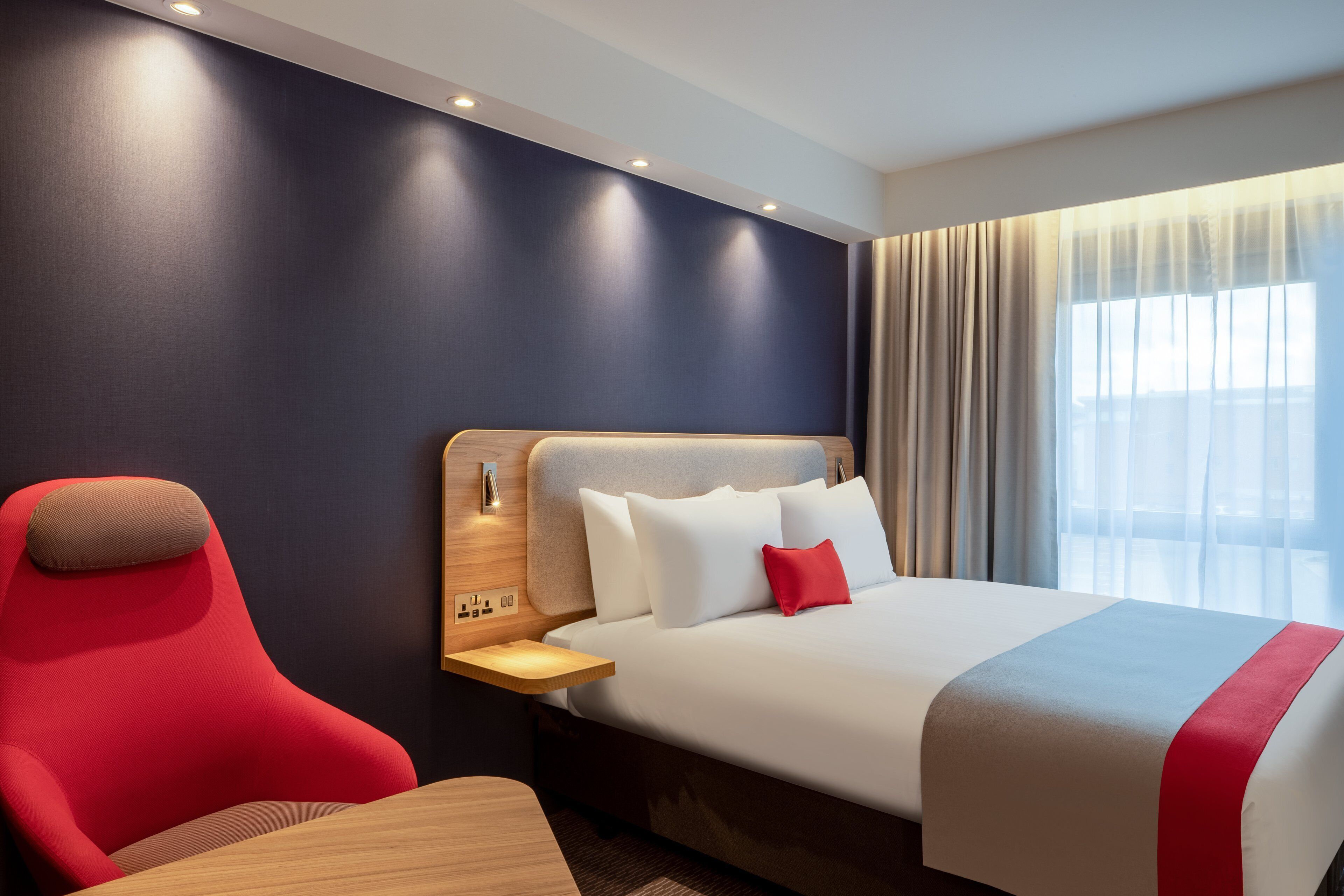 A hotel room with a bed, a red chair, a wooden nightstand, and a window with curtains.