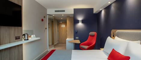 A hotel room with a bed, a red chair, a TV, a shelf with a kettle, and a small table.