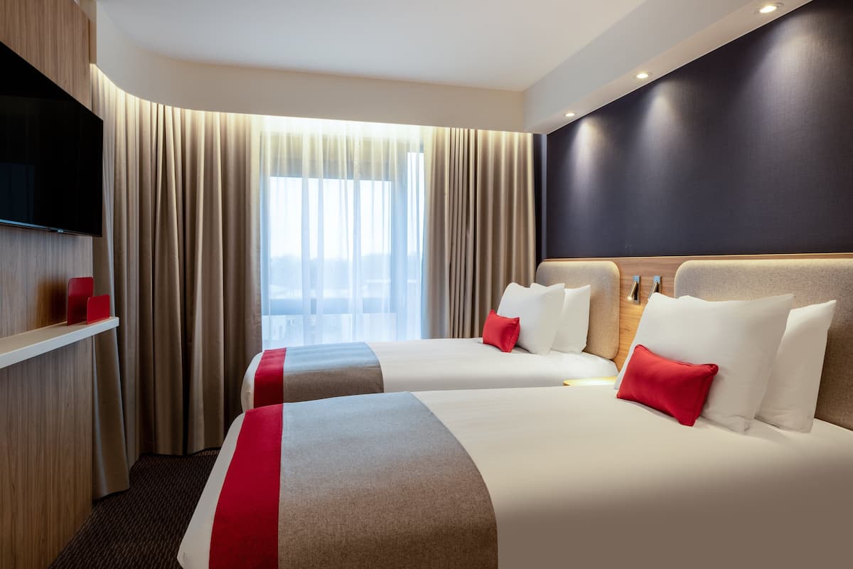 A hotel room with two beds, a flat-screen TV mounted on the wall, and a large window with curtains.