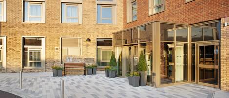 A modern building with a brick facade, large windows, and a paved entrance area with planters.