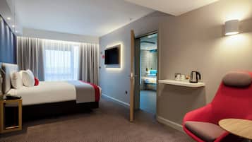 A hotel room with a bed, a red chair, a small table, a TV mounted on the wall, and a bathroom visible through an open door.