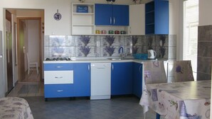 Uma cozinha compacta com armários azuis e brancos, painel de azulejos, pia e um espaço de jantar com mesa coberta por toalha de mesa florida.