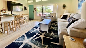 A spacious living area with a dining table, bar seating, a sofa, and a large window offering a view of the outdoors. - Well Appointed Oasis in the Sun! ☀️⚓️🏖 Pool, Beach, Boat Launch, Gorge, Wineries (Quincy)