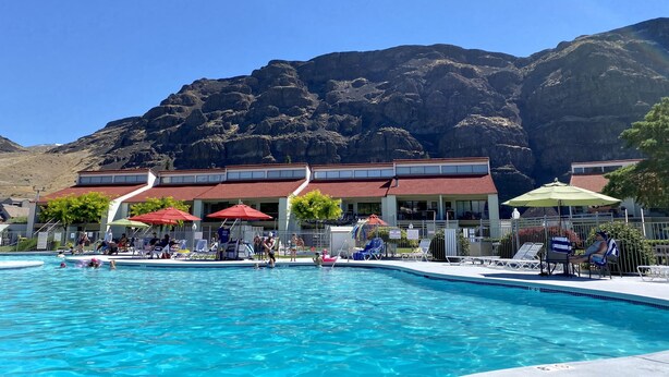 A swimming pool with lounge chairs and umbrellas, surrounded by a building with a red roof, set against a mountainous backdrop. - Well Appointed Oasis in the Sun! ☀️⚓️🏖 Pool, Beach, Boat Launch, Gorge, Wineries (Quincy)