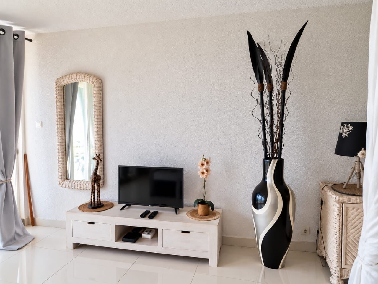 A modern living room with a flat-screen TV, a white console table, a tall vase with branches, and a mirror on the wall.