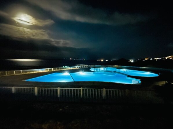 Vista nocturna de un complejo de piscinas con varias albercas y una valla alrededor.