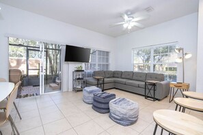 Un salon moderne avec un canapé d’angle, des poufs, un téléviseur à écran plat et un ventilateur de plafond.
