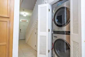 Une buanderie équipée de deux machines à laver à chargement frontal superposées, d’une porte blanche et d’un couloir menant à une pièce dotée d’une porte et d’un luminaire.