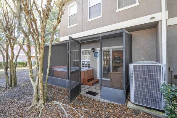 A covered outdoor seating area with a hot tub, a sliding glass door leading to a kitchen, and an air conditioning unit.