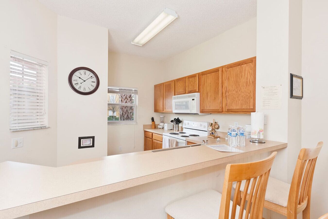 A modern kitchen with wooden cabinets, a white countertop, a stove, and a microwave. There is a clock on the wall and a window with blinds.