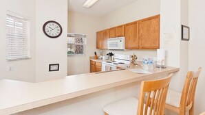 A modern kitchen with wooden cabinets, a white countertop, a stove, and a microwave. There is a clock on the wall and a window with blinds. - Emerald Island Townhome just 10min to Disney (Kissimmee)