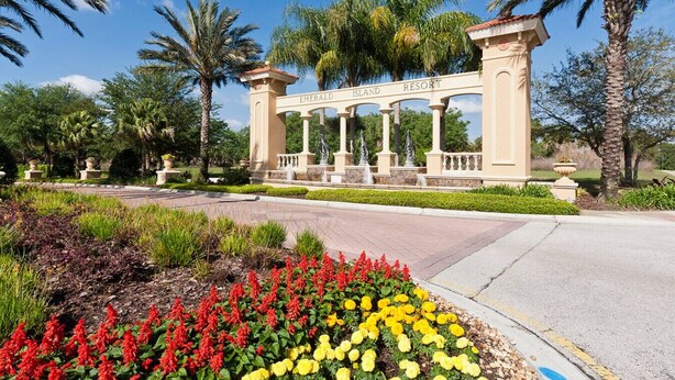 A gated entrance with a sign reading 'Emerald Island Resort,' surrounded by landscaped gardens with red and yellow flowers, palm trees, and a clear blue sky. - Emerald Island Townhome just 10min to Disney (Kissimmee)