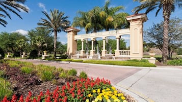 Una entrada con puerta adornada con un cartel que dice 'Emerald Island Resort', rodeada de jardines cuidados con flores rojas y amarillas, palmeras y un cielo azul despejado.