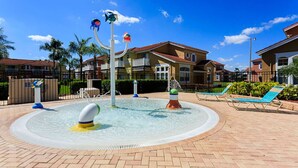 Un área de juegos acuáticos con una piscina circular, sillas de descanso y un edificio con techo rojo al fondo.