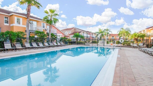 Una piscina con sillas de descanso, palmeras y una hilera de edificios al fondo.