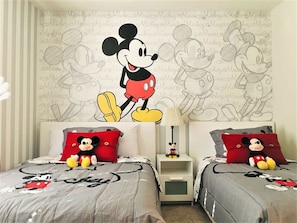 Quarto com duas camas, cada uma com roupa de cama temática do Mickey Mouse. Há almofadas do Mickey Mouse e um brinquedo de pelúcia do Mickey Mouse na mesinha de cabeceira.