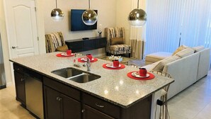 A modern kitchen with a granite countertop, stainless steel sink, and dark cabinetry. There is a dining area with a patterned sofa and a television mounted on the wall. - SpaciousTownhome 15 min to Disney Private Pool-BBQ (Kissimmee)