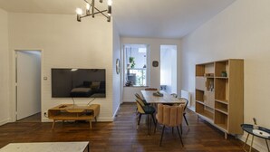 Un salon moderne avec un téléviseur à écran plat fixé au mur, une table basse en bois, un coin repas avec une table et des chaises, et une bibliothèque intégrée.