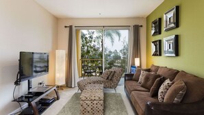 A living room with a flat-screen TV, a sofa, an ottoman, and a dining area with chairs.