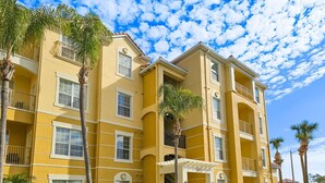 A yellow multi-story building with balconies, a central entrance, and palm trees. - Modern Condo  5min to Conv Center  with balcony (Orlando)