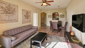 A living room with a sofa, coffee table, dining table with chairs, a ceiling fan, and a TV. - Modern Condo  5min to Conv Center  with balcony (Orlando)