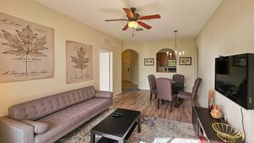 A living room with a sofa, coffee table, dining table with chairs, a ceiling fan, and a TV.