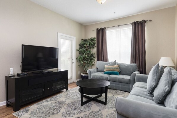 A living room with a flat-screen TV on a black stand, a grey sofa with cushions, a round coffee table, and a potted plant. - Spacious Condo 5min to Universal and Conv Center (Orlando)