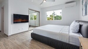 A modern bedroom with a large bed, a ceiling fan, a TV, and a window with a view of greenery. - The Makai Manor - Beautiful Newly Built Spacious Home (Honolulu)