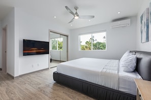 Ein modernes Schlafzimmer mit einem großen Bett, einem Deckenventilator, einem Fernseher und einem Fenster mit Blick auf Grünflächen.