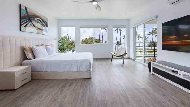 A modern bedroom with a large bed, a ceiling fan, a TV, and a view of the beach. - The Makai Manor - Beautiful Newly Built Spacious Home (Honolulu)