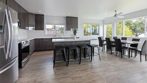 A modern kitchen with a central island, dark wood cabinets, and a large window providing a view of greenery outside. - The Makai Manor - Beautiful Newly Built Spacious Home (Honolulu)