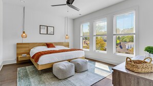 Une chambre à coucher moderne avec un lit en bois, deux poufs, un ventilateur de plafond et de grandes fenêtres.