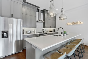Une cuisine moderne avec des meubles gris, un réfrigérateur en acier inoxydable, un îlot central blanc avec des tabourets de bar et un panneau lumineux au néon sur lequel on peut lire « Nashville ».