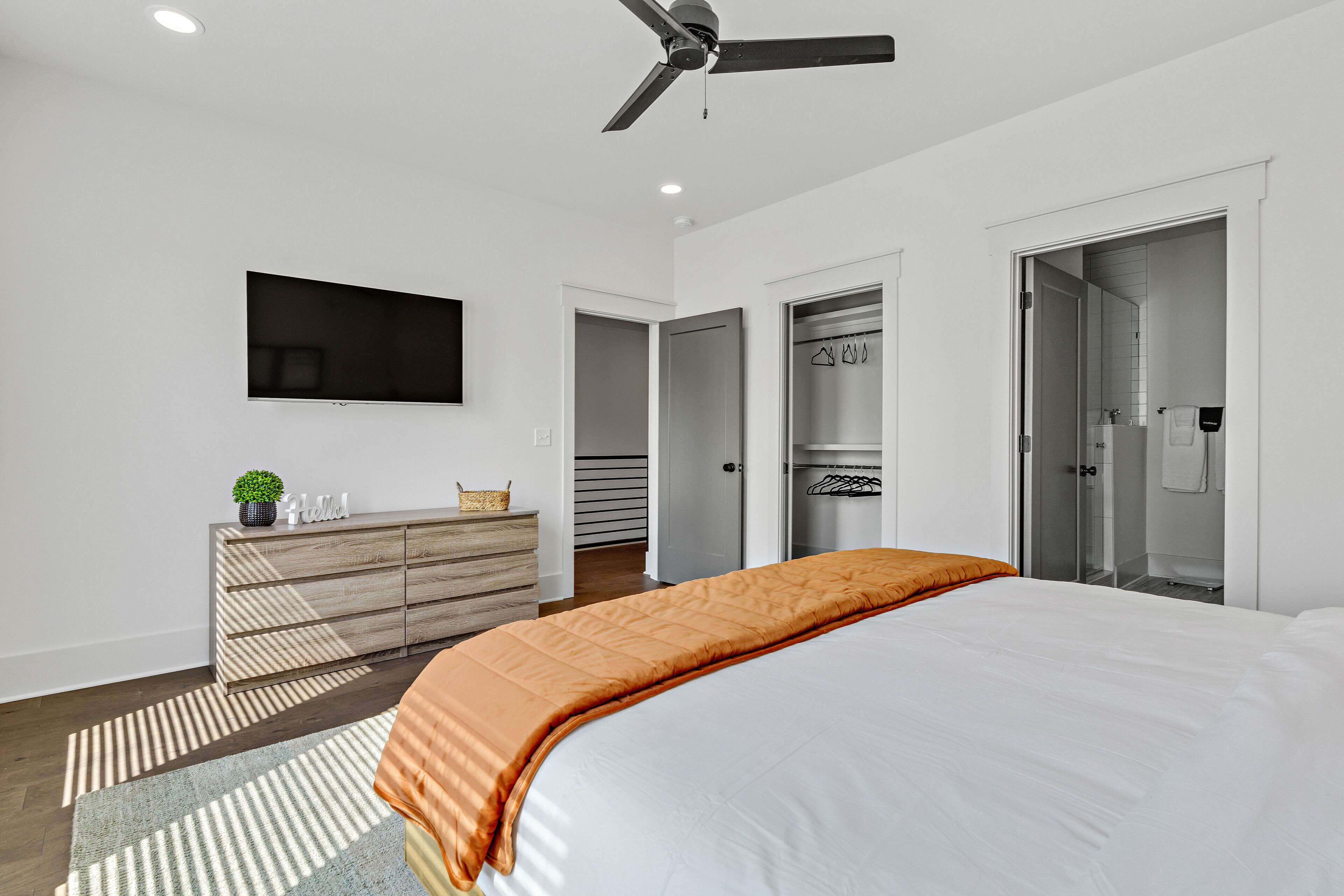 Ein modernes Schlafzimmer mit Bett, Fernseher, Deckenventilator, Kleiderschrank und angrenzendem Badezimmer.