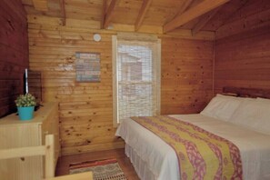 A wooden cabin bedroom with a bed, a window with blinds, a small table with a plant, and a wall-mounted sign.